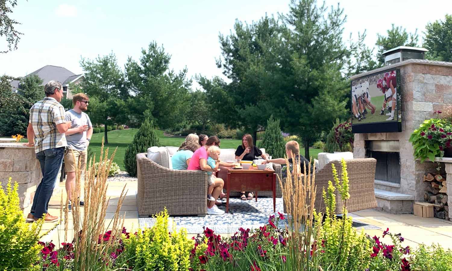 A group of people gathered in a landscaped backyard, enjoying a social gathering with outdoor seating, a fireplace, and a TV displaying a football game.