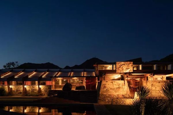 A modern architectural building illuminated at night with strategically placed outdoor lighting, highlighting the stone textures, wooden elements, and structural details against a mountainous backdrop. The warm lighting creates a welcoming ambiance while emphasizing the building's design features.