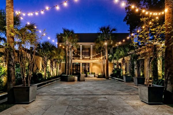 Illuminated courtyard at dusk with string lights overhead, surrounding palm trees, and a modern building as the backdrop.