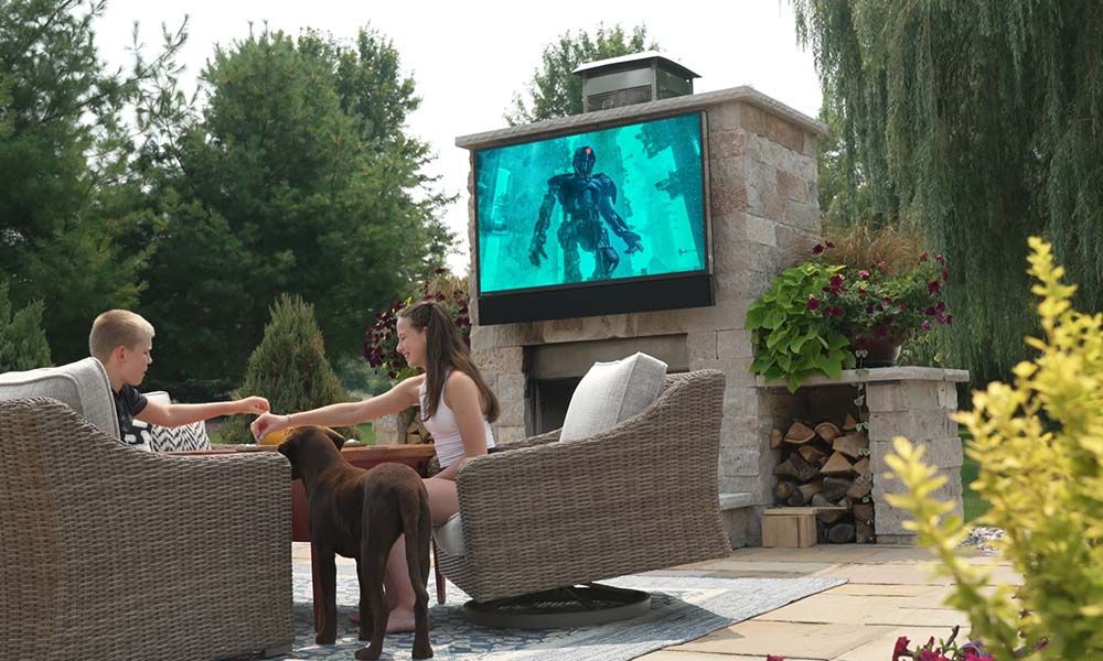 Two children sitting on outdoor wicker chairs with a dog nearby, watching a sci-fi movie on a large TV mounted above a stone fireplace in a landscaped backyard.